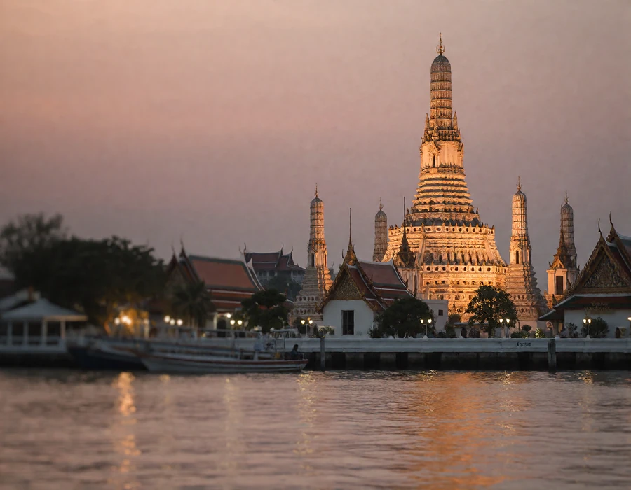 Riverside view of an illuminated Thai temple spire against a dusky sky reflected in calm water